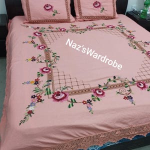 May include: A pink bedspread and two pillowcases with embroidered floral designs. The bedspread features a large square border with a floral pattern and the text "Naz'sWardrobe". The pillowcases have a single rose design.