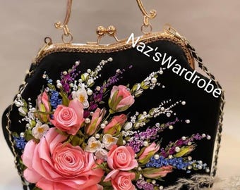 May include: Black handbag with a gold handle and clasp, adorned with a large floral arrangement of pink roses, white pearls, and other colorful flowers. The text "Naz's Wardrobe" is visible on the bag.