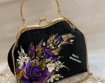 May include: Black velvet handbag with a gold chain strap and handle. The bag is adorned with a floral arrangement of purple, white, and yellow ribbon roses, wheat, and pearls. The text "Naz's Wardrobe" is printed on the side.