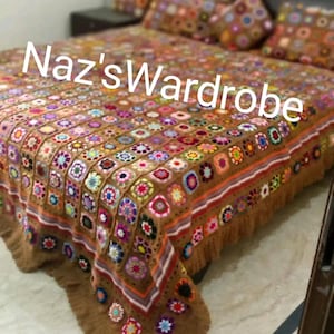 May include: A handmade, brown crochet bedspread with a colorful floral pattern. The bedspread features a patchwork design with various colored floral squares and a decorative border. The text "Naz's Wardrobe" is across the top of the bedspread.