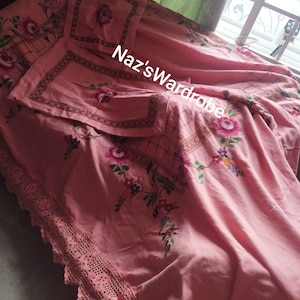 May include: A pink floral bedspread with matching pillowcases. The bedspread features embroidered roses and a decorative border. The edge of the bedspread has a crocheted lace trim. The text "Naz's Wardrobe" is visible on the bedspread.