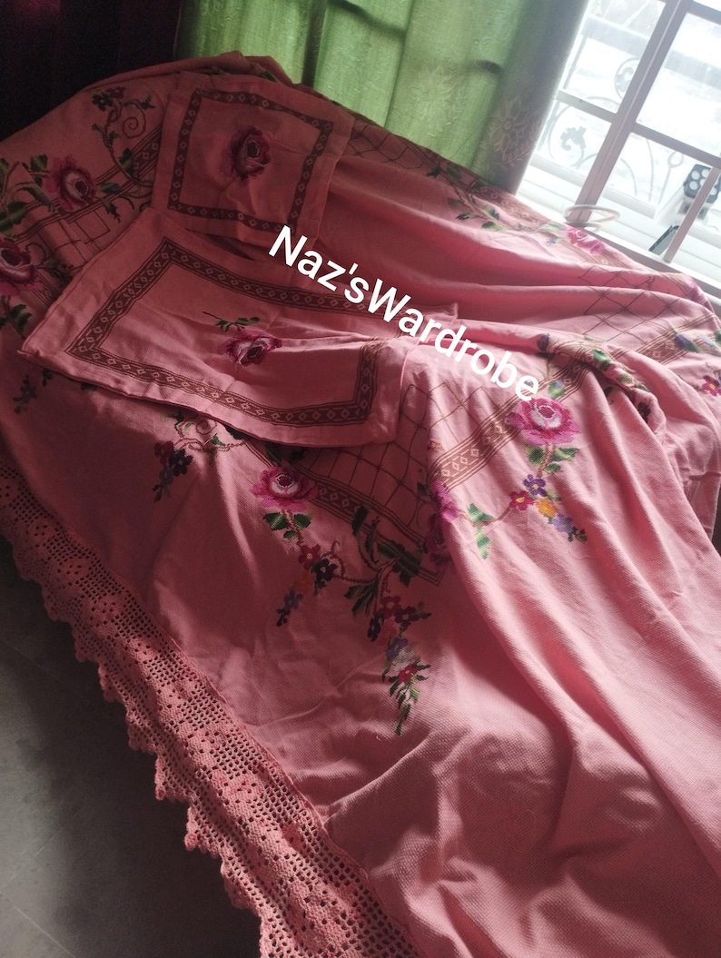 May include: A pink floral bedspread with matching pillowcases. The bedspread features embroidered roses and a decorative border. The edge of the bedspread has a crocheted lace trim. The text "Naz's Wardrobe" is visible on the bedspread.