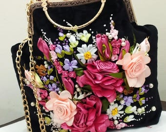 May include: A black velvet handbag with a gold chain strap and a floral design embroidered with pink, purple, and white ribbon. The bag has a gold clasp and a bow with rhinestones.