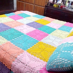 May include: A handmade, colorful crochet blanket with a patchwork design. The blanket features squares in pastel shades of pink, yellow, blue, and peach. Two matching crochet pillows are also visible, adding a decorative touch to the bed.
