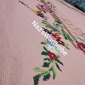May include: Close-up of a pink cross-stitch fabric with a floral design. The embroidery features red, pink, yellow, blue, and white flowers with green leaves. The text "Naz's Wardrobe" is visible in white.
