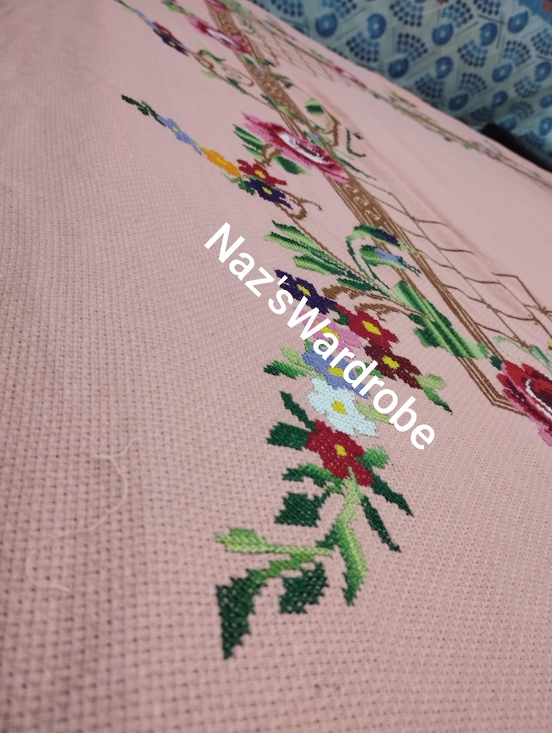 May include: Close-up of a pink cross-stitch fabric with a floral design. The embroidery features red, pink, yellow, blue, and white flowers with green leaves. The text "Naz's Wardrobe" is visible in white.