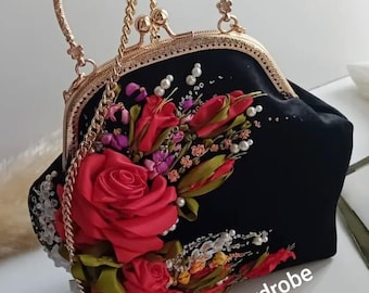 May include: Black handbag with a gold-tone frame, handle, and chain strap. The bag is adorned with red ribbon roses, white pearls, and other colorful floral accents. The text "Naz's Wardrobe" is visible on the bag.