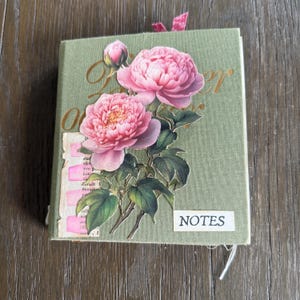 Könnte beinhalten: Ein salbeigrünes, stoffbezogenes Notizbuch mit einer großen Blumen-Collage aus rosa Pfingstrosen und grünen Blättern auf dem Einband. Ein kleines weißes rechteckiges Etikett mit dem schwarzen Text "NOTES" ist zusammen mit altem Papier angebracht.