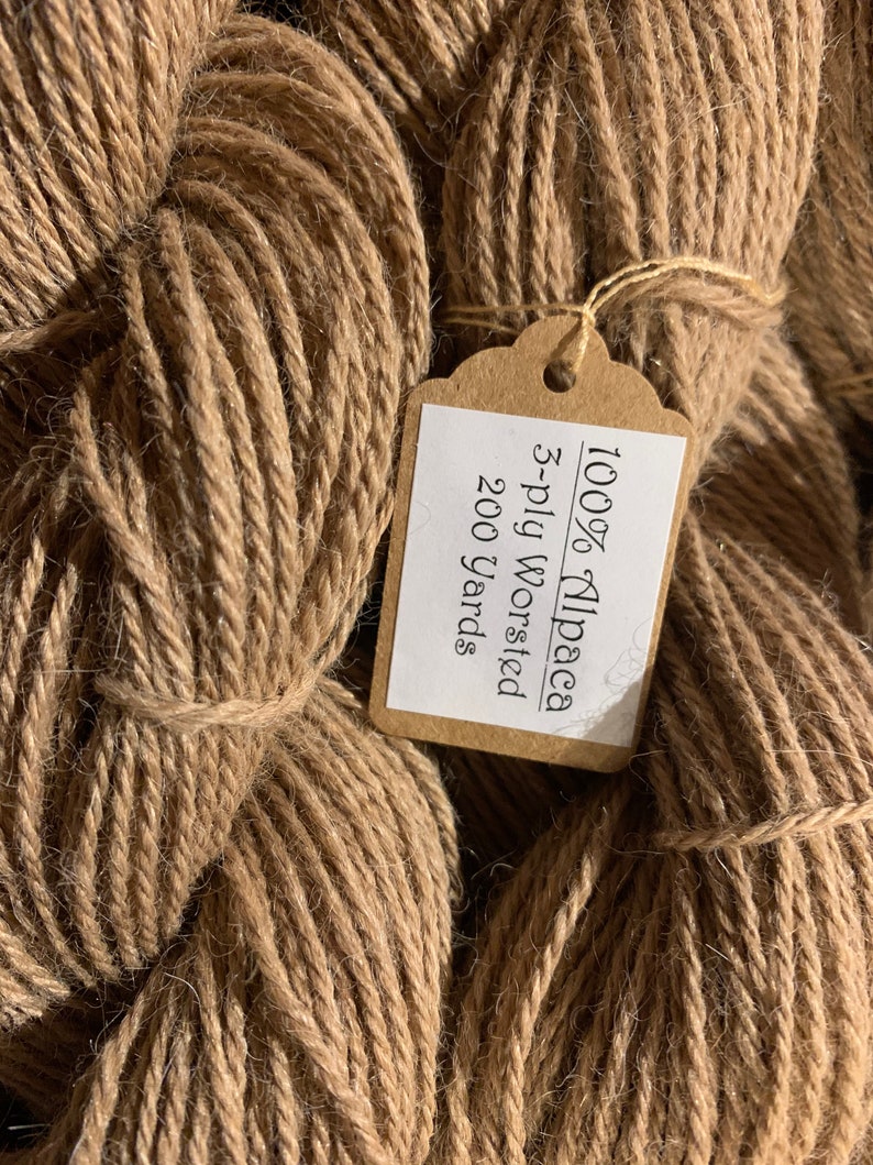 Puede incluir: Una madeja de hilo de alpaca marr&oacute;n con una etiqueta que dice "100% Alpaca 3-ply worsted 200 yards".