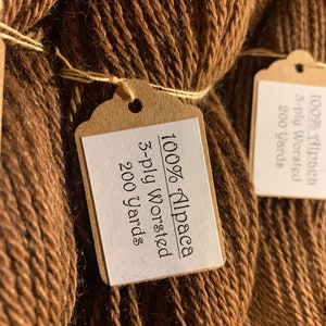 Puede incluir: Dos ovillos de hilo de alpaca marr&oacute;n al 100%, cada uno con una etiqueta de papel marr&oacute;n que dice "100% Alpaca 3-ply Worsted 200 yards".