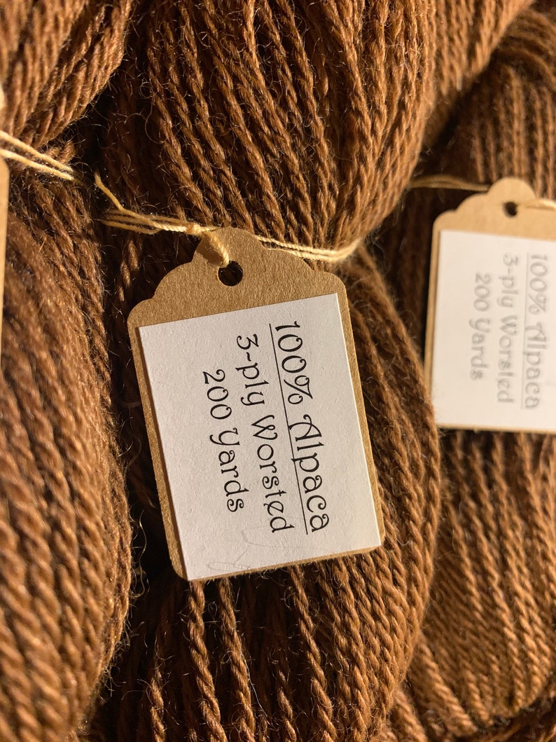 Puede incluir: Dos ovillos de hilo de alpaca marr&oacute;n al 100%, cada uno con una etiqueta de papel marr&oacute;n que dice "100% Alpaca 3-ply Worsted 200 yards".
