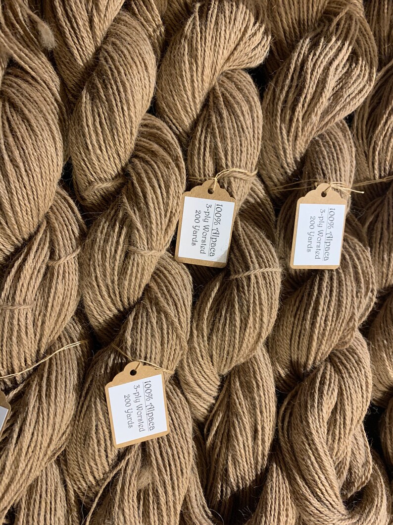 Puede incluir: Primer plano de varios carretes de hilo de alpaca marr&oacute;n con etiquetas de papel que dicen "100% Alpaca 3-ply Worsted 200 Yards".