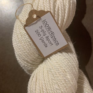 Puede incluir: Una madeja de hilo blanco de alpaca 100%, etiquetada como hilo deportivo de 3 cabos con 200 yardas.