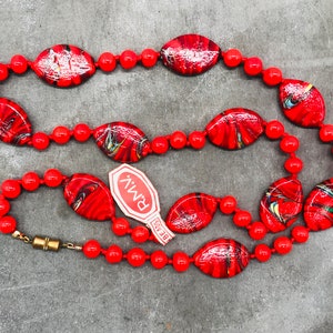 K&ouml;nnte beinhalten: Eine Halskette aus roten und schwarzen Perlen aus Glas mit einem goldenen Verschluss. Die Halskette liegt auf einer grauen Oberfl&auml;che mit einem wei&szlig;en Etikett, auf dem "BEADS" und "PMIV" steht.