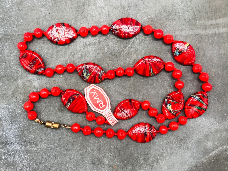 K&ouml;nnte beinhalten: Eine Halskette aus roten und schwarzen Perlen aus Glas mit einem goldenen Verschluss. Die Halskette liegt auf einer grauen Oberfl&auml;che mit einem wei&szlig;en Etikett, auf dem "BEADS" und "PMIV" steht.