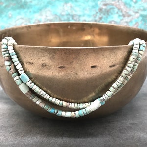 K&ouml;nnte beinhalten: Zwei Str&auml;nge t&uuml;rkisfarbener Perlen in einem hellblauen und wei&szlig;en Farbschema werden in einer Messing Schale pr&auml;sentiert. Die Perlen sind klein und quadratisch.