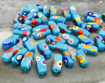 Puede incluir: Una colección de cuentas de vidrio azul con patrones coloridos, multicolores y blancos. Las cuentas son de forma rectangular y tienen un acabado liso y brillante.