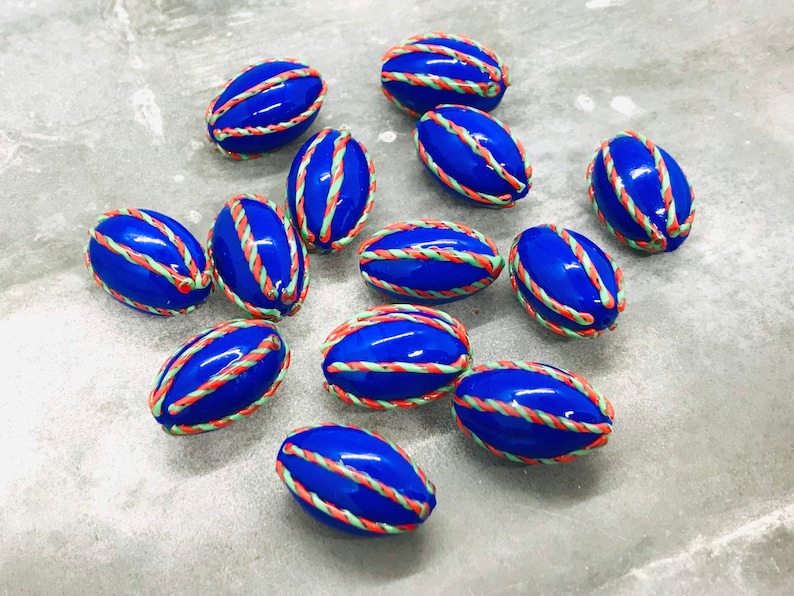 Op de afbeelding: Een verzameling van twaalf blauwe, ovale kralen met witte en oranje randen. De kralen zijn gerangschikt op een grijs oppervlak.
