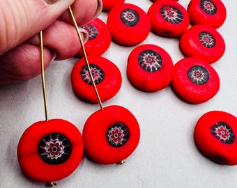 Puede incluir: Cuentas redondas de color rojo brillante con un diseño floral en el centro. Se están ensartando las cuentas en alambres dorados para crear pendientes. El fondo es de color gris claro.