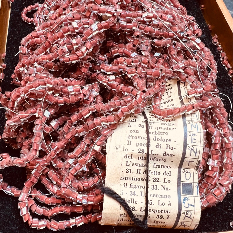 May include: A pile of translucent, pink, square-shaped beads strung together on a thin wire. The beads are arranged in a random pattern and are partially covering a piece of paper with text in Italian.
