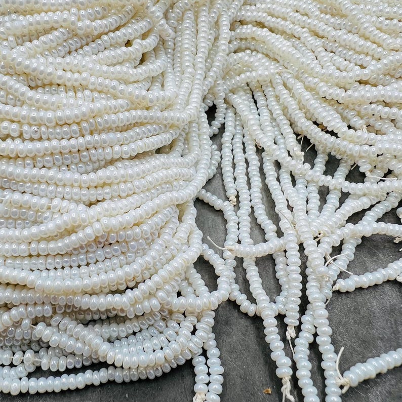May include: A large quantity of white seed beads, also known as rocailles, are piled together. The beads are small and round, with a slightly iridescent finish.