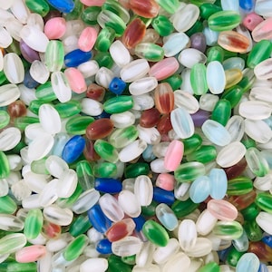 Pu&ograve; includere: Un vasto assortimento di perline di vetro ovali in vari colori, tra cui trasparente, verde, blu, rosa e marrone. Le perline sono traslucide e hanno un leggero bagliore iridescente.