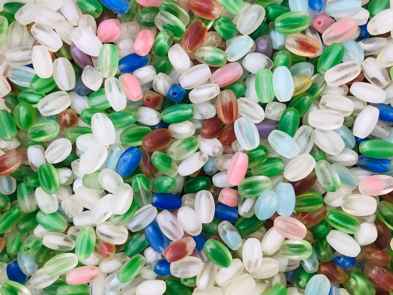 Pu&ograve; includere: Un vasto assortimento di perline di vetro ovali in vari colori, tra cui trasparente, verde, blu, rosa e marrone. Le perline sono traslucide e hanno un leggero bagliore iridescente.