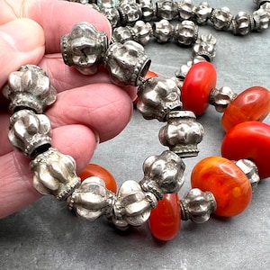 May include: A close-up of a silver and orange beaded necklace. The necklace is made of small, round silver beads with a textured surface and larger, round orange beads. The beads are strung together on a silver chain.