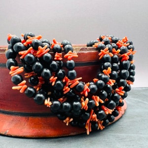 Puede incluir: Un collar de múltiples hilos hecho de cuentas negras y detalles de color coral. El collar está drapeado sobre una caja de madera.