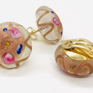 May include: A pair of gold clip-on earrings with round, clear glass stones. The stones are decorated with pink, blue, and gold swirls and dots.