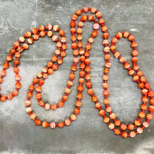 May include: A long necklace made of orange and white striped beads. The beads are strung on a thin cord and the necklace is lying on a gray surface.