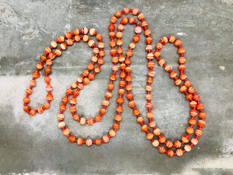 May include: A long necklace made of orange and white striped beads. The beads are strung on a thin cord and the necklace is lying on a gray surface.