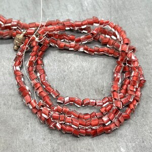 May include: A strand of red glass beads with a clear, iridescent finish. The beads are square shaped and have a slightly irregular shape. The beads are strung on a clear thread.
