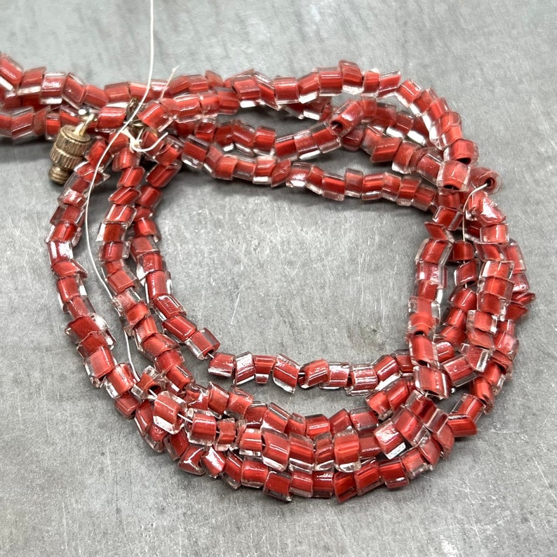 May include: A strand of red glass beads with a clear, iridescent finish. The beads are square shaped and have a slightly irregular shape. The beads are strung on a clear thread.