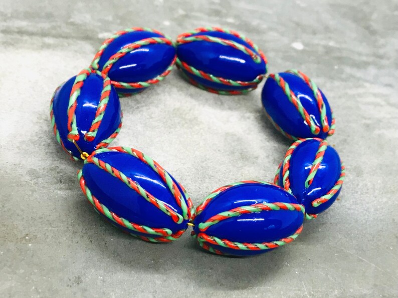 Op de afbeelding: Een armband gemaakt van blauwe, groene en oranje ovale kralen. De kralen zijn versierd met een touwachtig patroon.