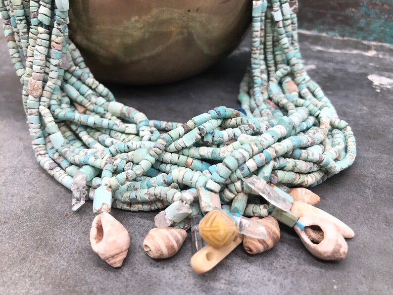 K&ouml;nnte beinhalten: Eine t&uuml;rkisfarbene und wei&szlig;e Perlenkette mit Muschel- und Kristall-Akzenten. Die Kette besteht aus vielen Perlenstr&auml;ngen und hat verschiedene Anh&auml;nger.