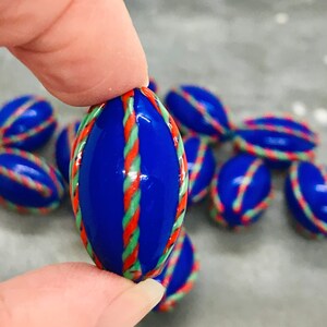 Op de afbeelding: Een groep blauwe glazen knikkers met rode, groene en witte strepen. De knikkers zijn ovaalvormig en hebben een glanzende afwerking.
