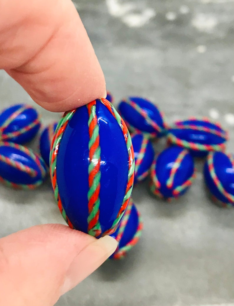 Op de afbeelding: Een groep blauwe glazen knikkers met rode, groene en witte strepen. De knikkers zijn ovaalvormig en hebben een glanzende afwerking.
