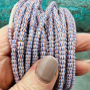 Pode incluir: Um close-up de um fio de contas de vidro listradas de branco e vermelho. As contas são pequenas e redondas, e são enfiadas em um fio branco.