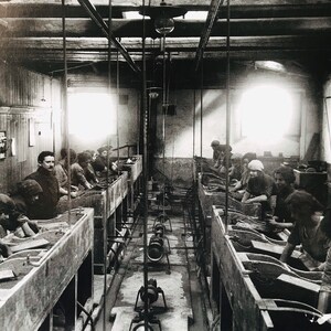 May include: Black and white photo of a factory setting with workers at long tables, likely processing or manufacturing something. The workers are seated and appear to be using tools or machinery.