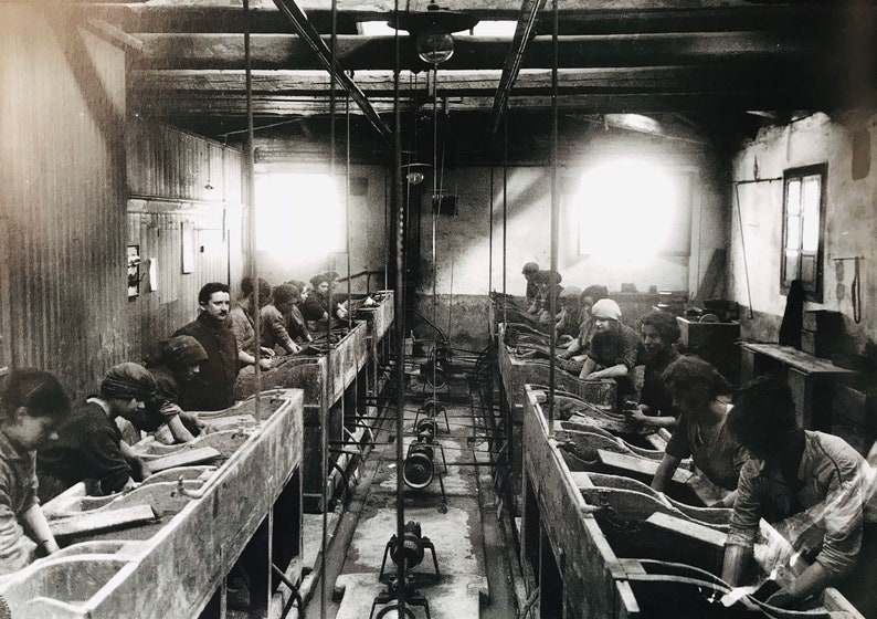 May include: Black and white photo of a factory setting with workers at long tables, likely processing or manufacturing something. The workers are seated and appear to be using tools or machinery.