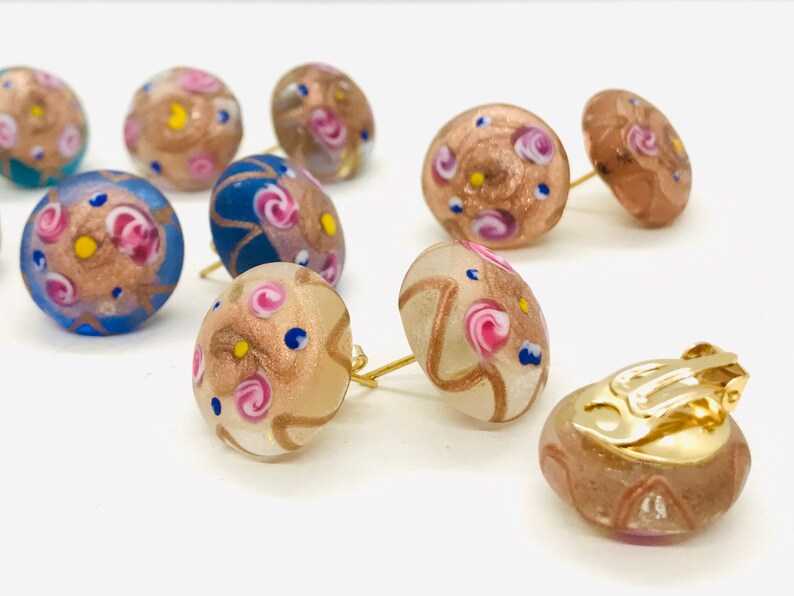 May include: A set of colorful glass earrings with a gold finish. The earrings feature a variety of patterns and colors, including pink, blue, and gold. The earrings are available in both stud and clip-on styles.