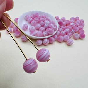 May include: Two pink and white striped glass beads on a gold wire. The beads are surrounded by a pile of similar beads.