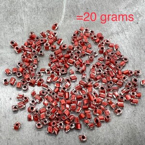 May include: A pile of approximately 20 grams of small, red, square-shaped glass beads. The beads have a clear, transparent finish with a red tint.