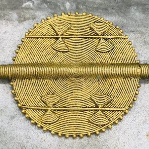 Può includere: Un ciondolo circolare in metallo dorato con un design a cerchi concentrici in rilievo. Il ciondolo ha una barra cilindrica che si estende dal centro del cerchio.