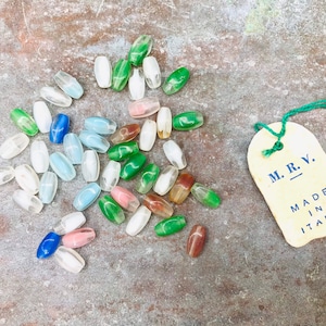 Pu&ograve; includere: Una collezione di perline di vetro colorate in varie tonalit&agrave; di blu, verde, rosa e bianco. Le perline sono di forma ovale e hanno una finitura liscia e lucida. Nell'immagine &egrave; visibile anche un piccolo cartellino con il testo "M.R.V. MADE IN ITALY".