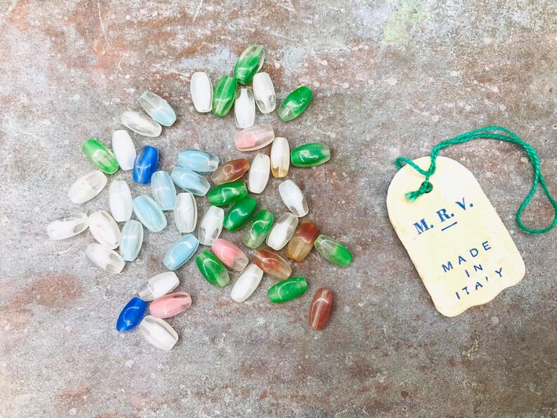 Pu&ograve; includere: Una collezione di perline di vetro colorate in varie tonalit&agrave; di blu, verde, rosa e bianco. Le perline sono di forma ovale e hanno una finitura liscia e lucida. Nell'immagine &egrave; visibile anche un piccolo cartellino con il testo "M.R.V. MADE IN ITALY".