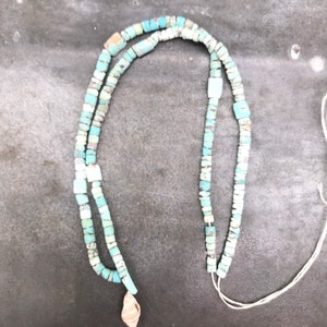 K&ouml;nnte beinhalten: Eine t&uuml;rkisfarbene Perlenkette mit einem kleinen Muschel-Anh&auml;nger. Die Kette besteht aus quadratischen Perlen und ist auf einer wei&szlig;en Schnur aufgef&auml;delt.