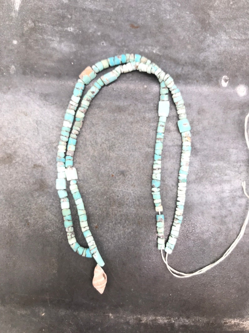 K&ouml;nnte beinhalten: Eine t&uuml;rkisfarbene Perlenkette mit einem kleinen Muschel-Anh&auml;nger. Die Kette besteht aus quadratischen Perlen und ist auf einer wei&szlig;en Schnur aufgef&auml;delt.