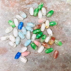 Pu&ograve; includere: Una collezione di perline di vetro ovali in vari colori, tra cui verde, blu, bianco e rosa. Le perline sono sparse su una superficie testurizzata.